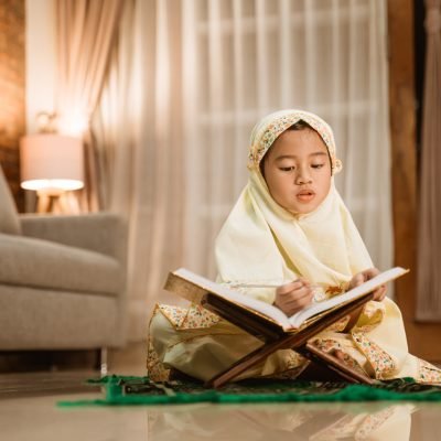 beautiful little girl reading quran wearing muslim hijab at home by herself
